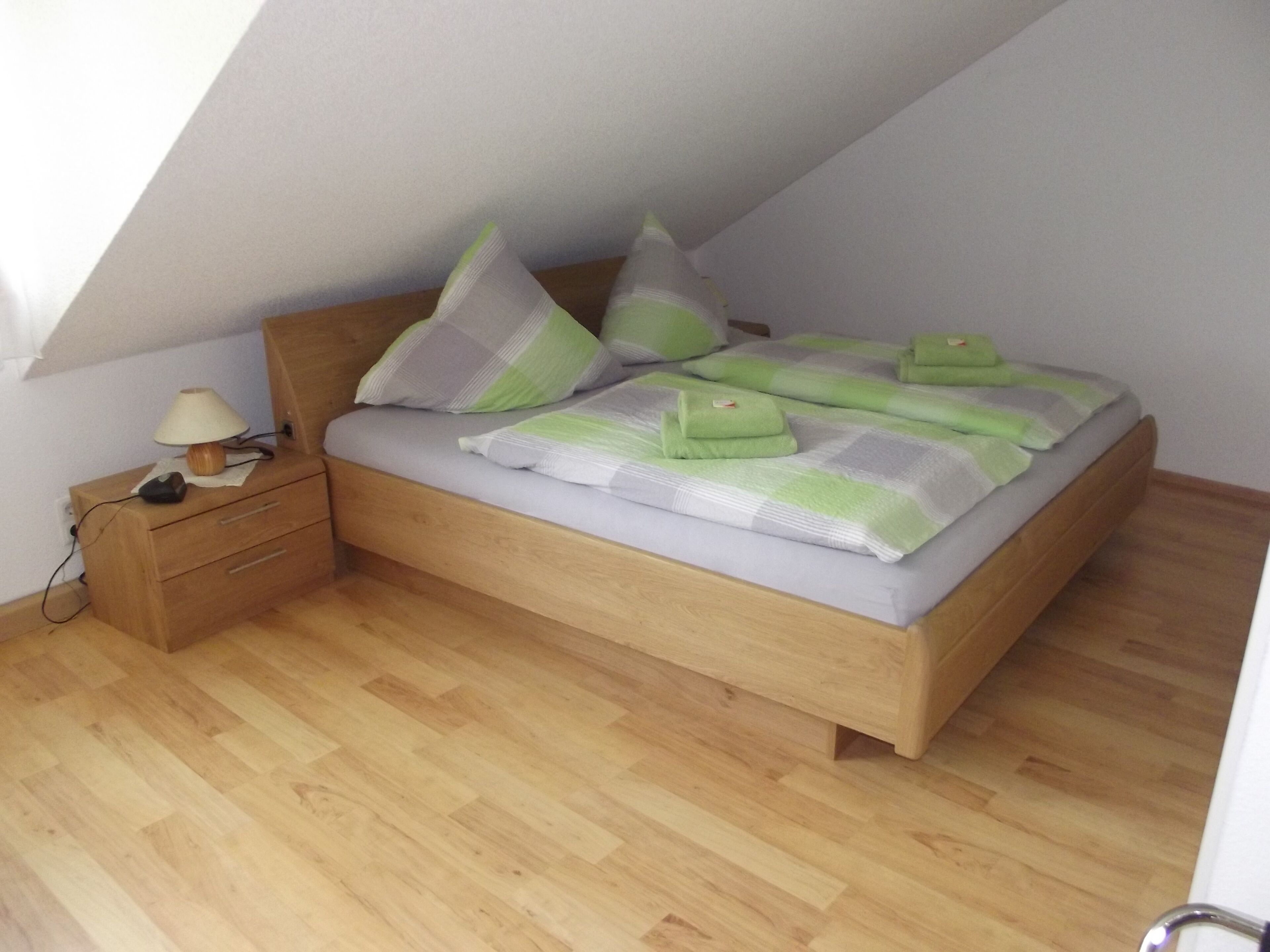 Een slaapkamer met een schuine kap, een houten bed met beddengoed in groene en grijze strepen, een nachtkastje van hout met een lamp, en een houten vloer.