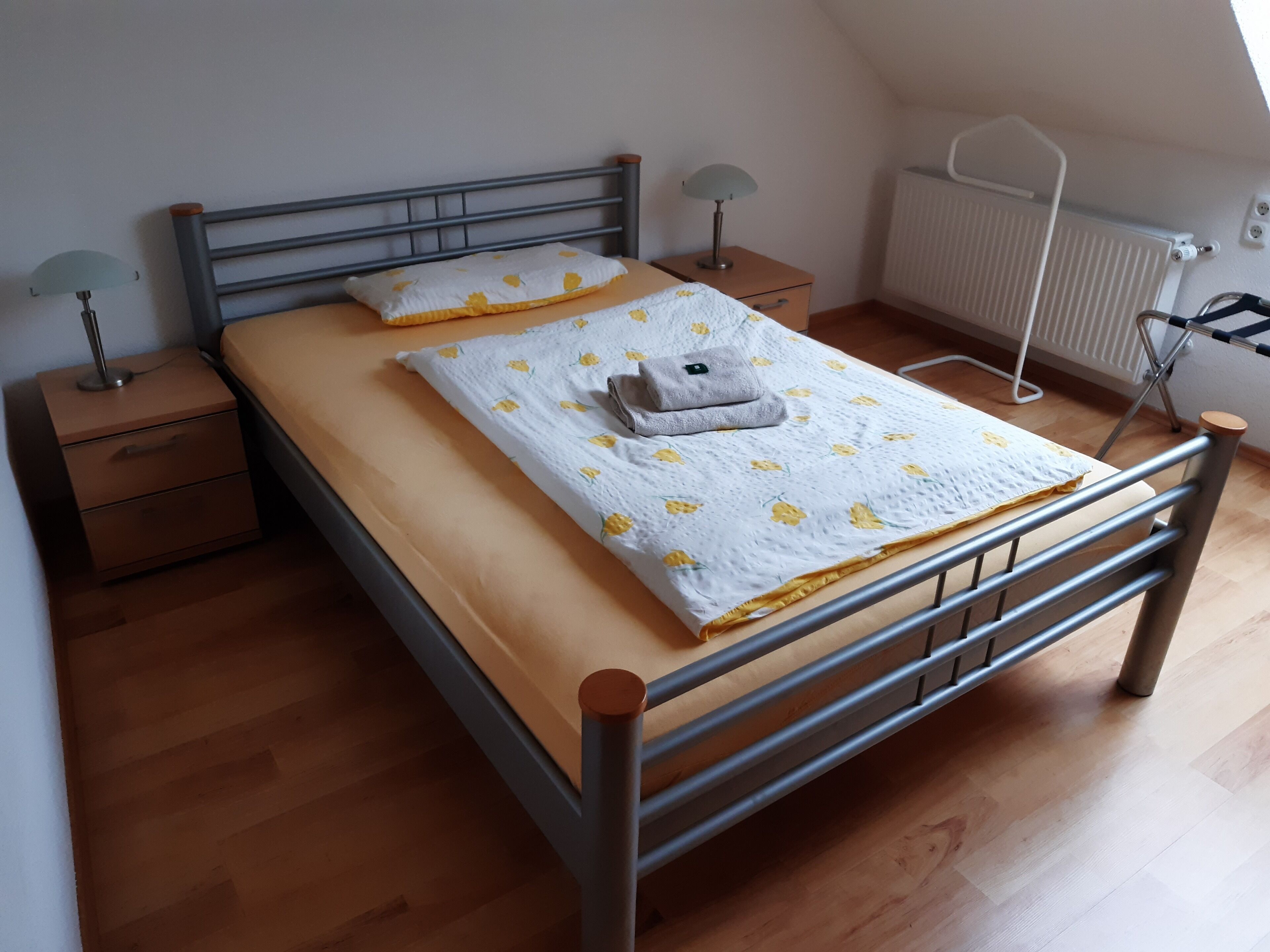 A single bed with a wooden headboard, a nightstand with a lamp, a radiator, and a window with a view of the sky.
