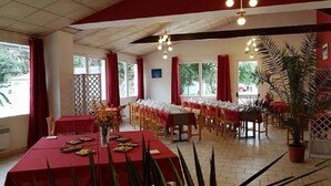 Un comedor con mesas dispuestas para una comida, sillas organizadas en filas y una planta grande en maceta en la esquina.