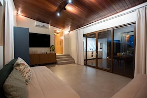 Una sala moderna con un sofá, un televisor de pantalla plana, techo de madera y una puerta corrediza de vidrio que da a una zona de cocina.