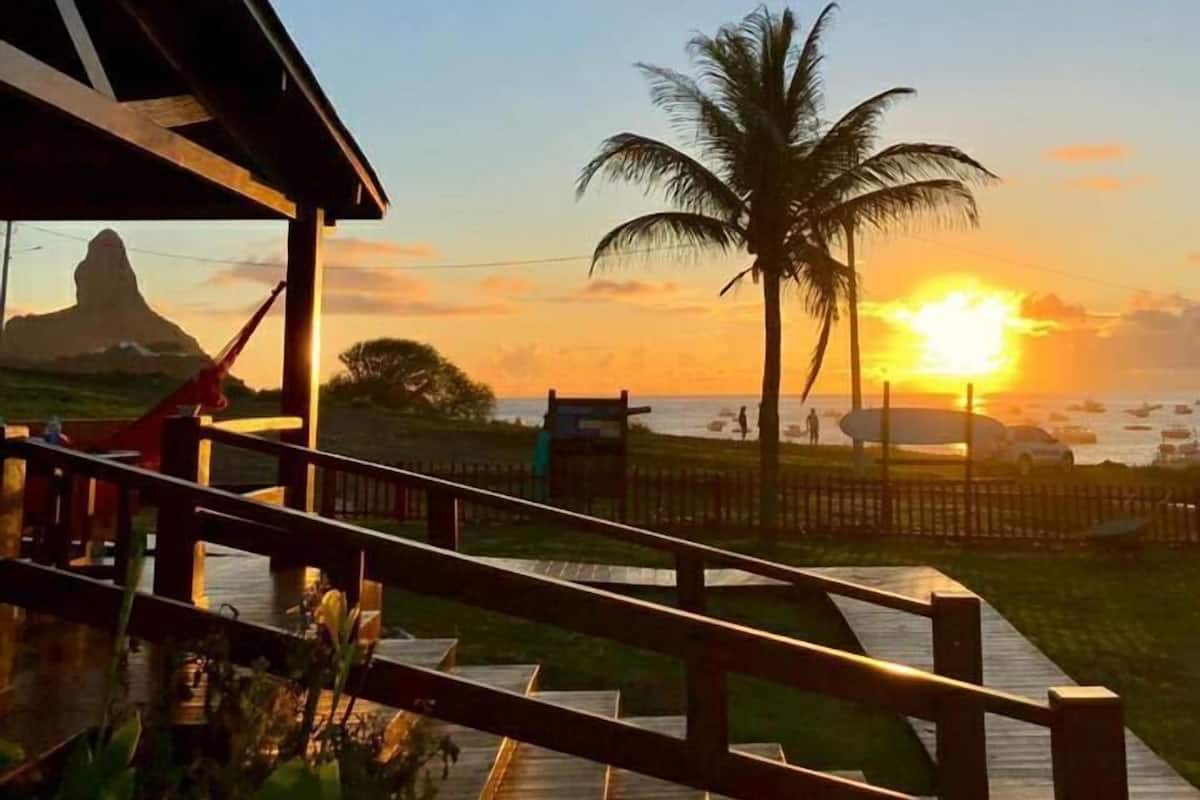 A wooden deck with a view of the sunset over the ocean, palm trees, and a mountain in the distance.