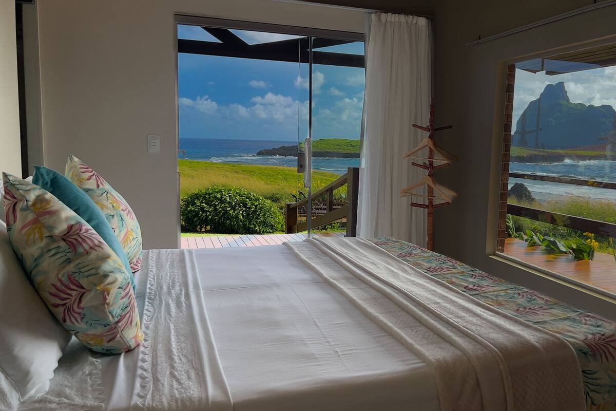 A bedroom with a bed, a view of the ocean, and a balcony with a wooden railing.