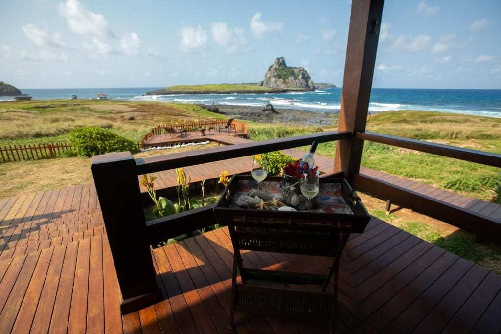 A wooden deck with a table set for dining, overlooking a coastal landscape with a prominent rock formation.
