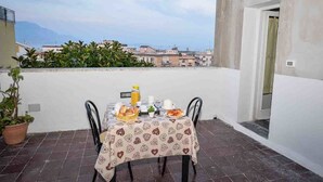 A table set on a balcony with breakfast items, a potted plant, and a view of buildings in the background.