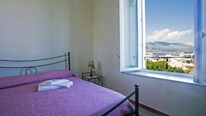 A bedroom with a bed, a nightstand, a lamp, and a window with a view of buildings and mountains.