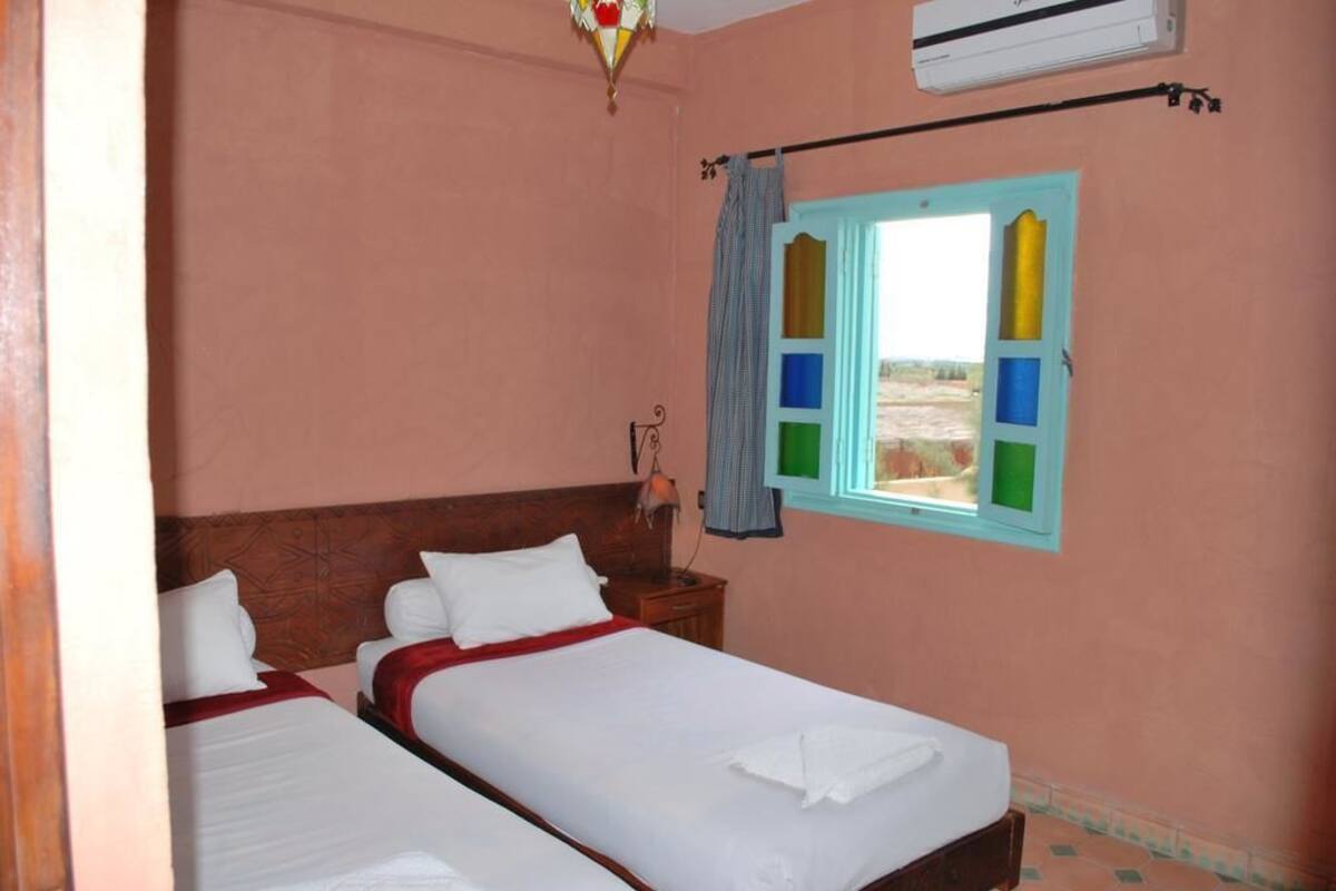 A room with two beds, a wooden headboard, a window with colorful panes, a hanging lamp, and an air conditioning unit.