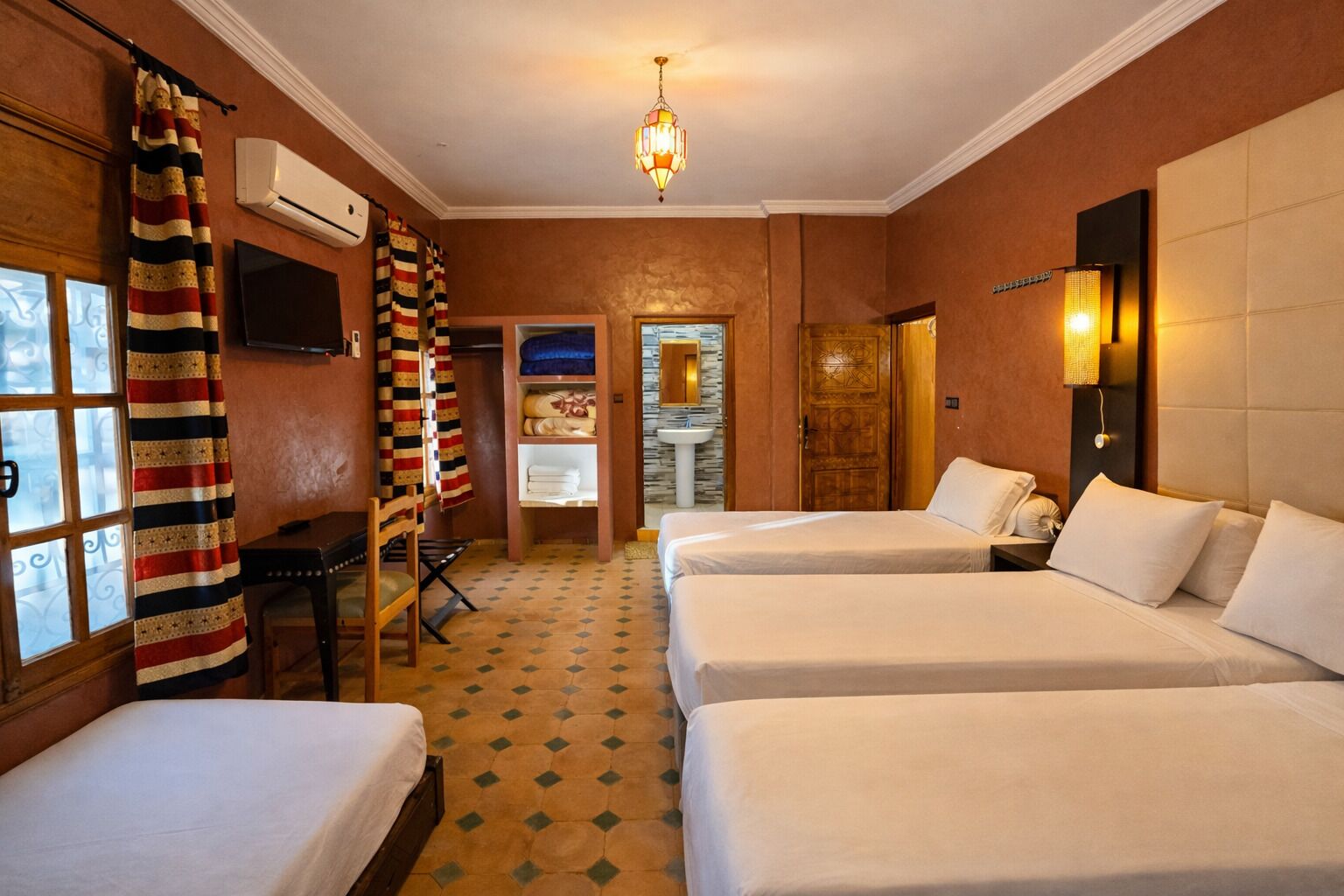 Une chambre d’hôtel avec trois lits, un bureau, une chaise, une télévision et une salle de bain visible à travers une porte ouverte.