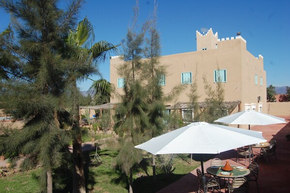 A building with a castle-like facade, outdoor seating with white umbrellas, and surrounding greenery.