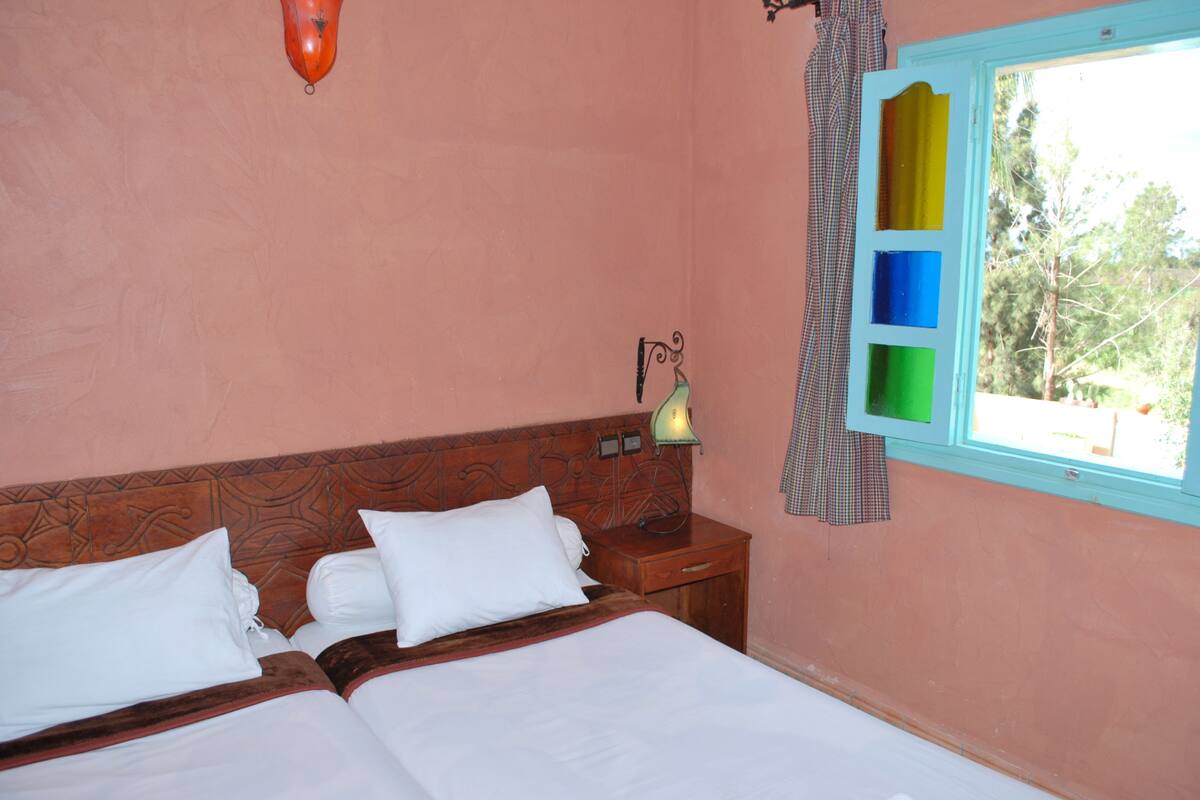 A bedroom with two beds, a nightstand with a lamp, and a window with curtains.