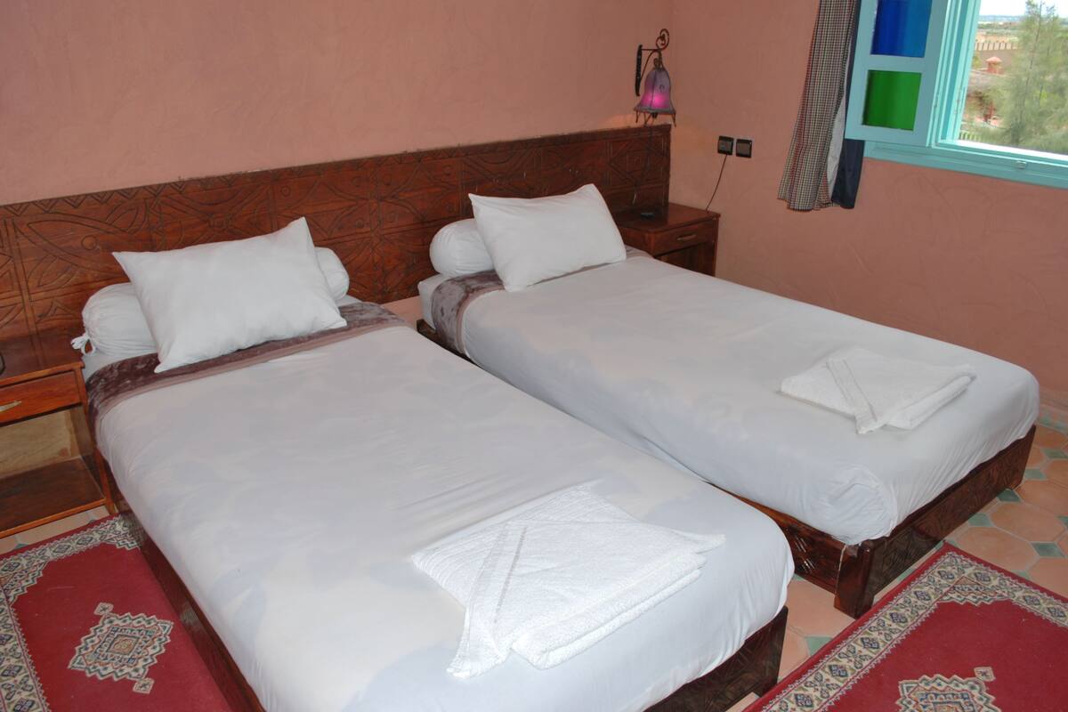 A double bed with white linens and a red patterned rug. There is a nightstand with a lamp and a telephone.