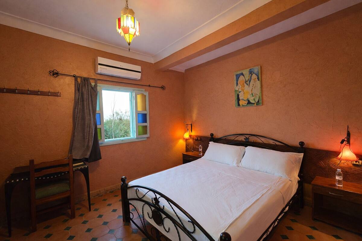 A bedroom with a bed, a window with a view, an air conditioning unit, and a chandelier.