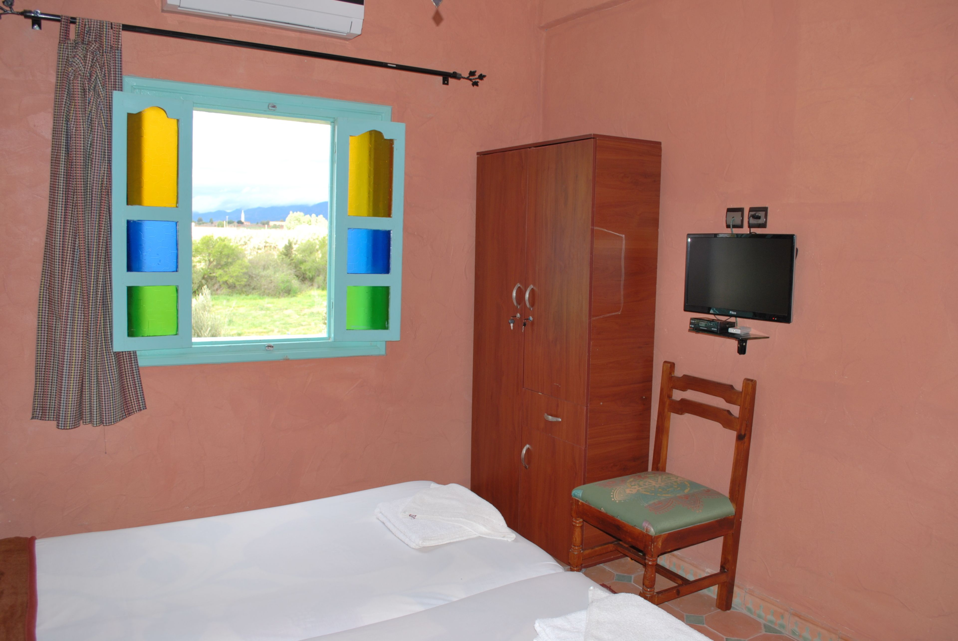 A bedroom with a bed, a wooden wardrobe, a television mounted on the wall, and a window with a view of greenery.