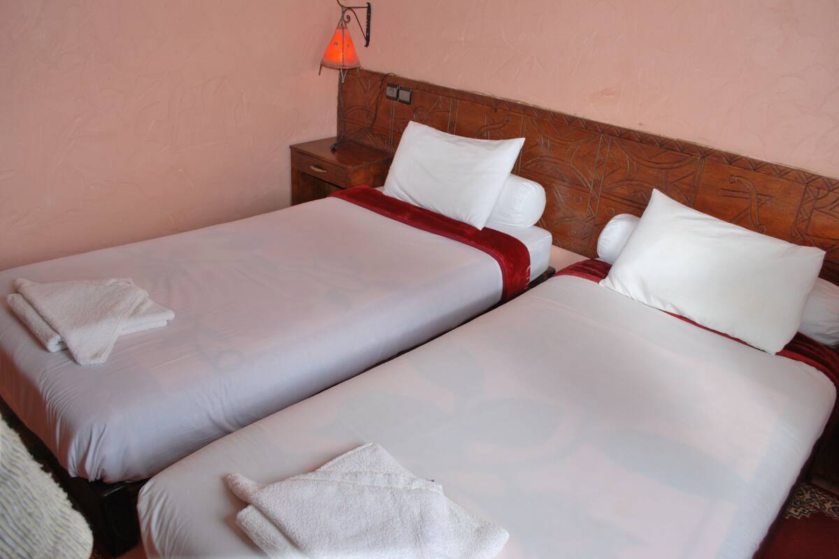 Two single beds with white linens and red accents, a wooden headboard, a nightstand with a lamp, and a pink wall.