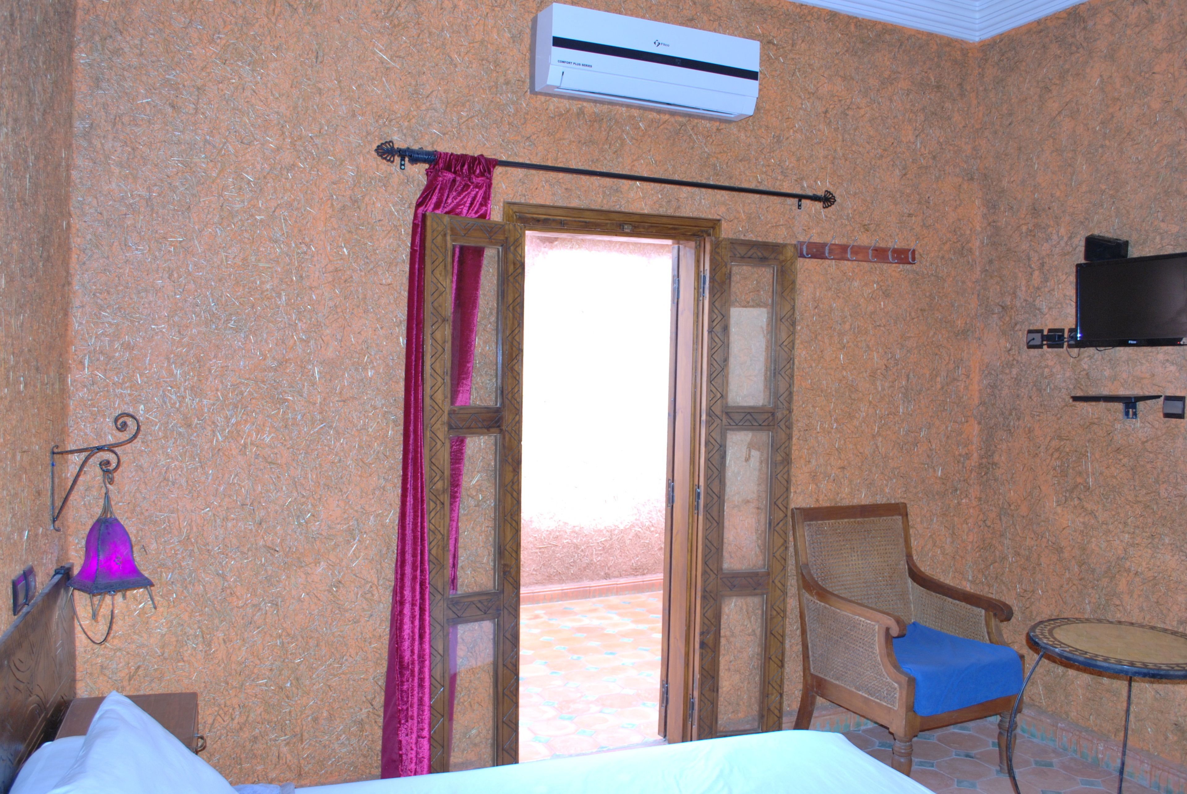 Habitación de hotel con una cama, una silla con cojín azul, una mesita y un equipo de aire acondicionado, además de vistas a través de una puerta abierta.