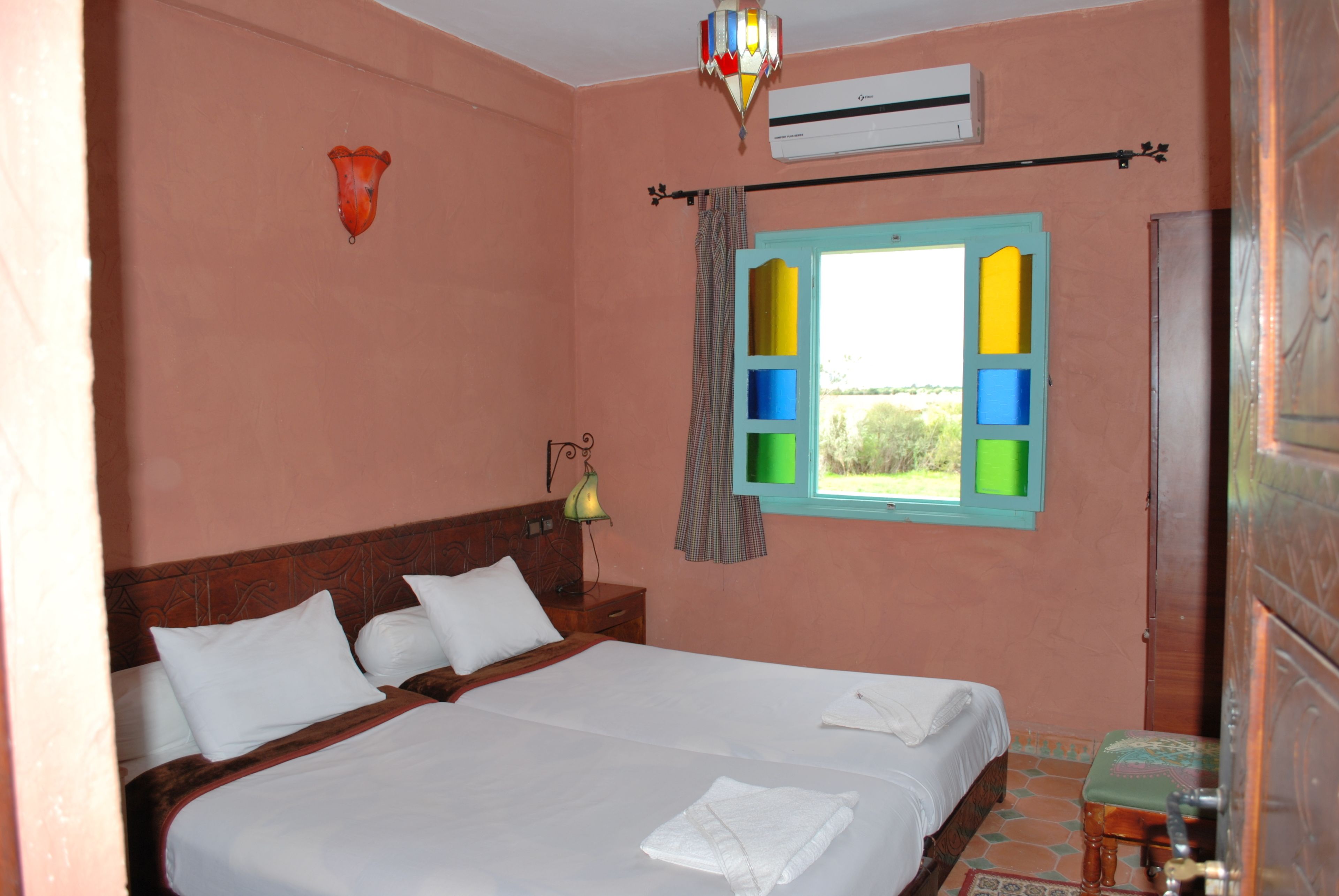 Une chambre avec deux lits, une fenêtre avec des volets colorés, un climatiseur fixé au mur et une lampe de chevet.