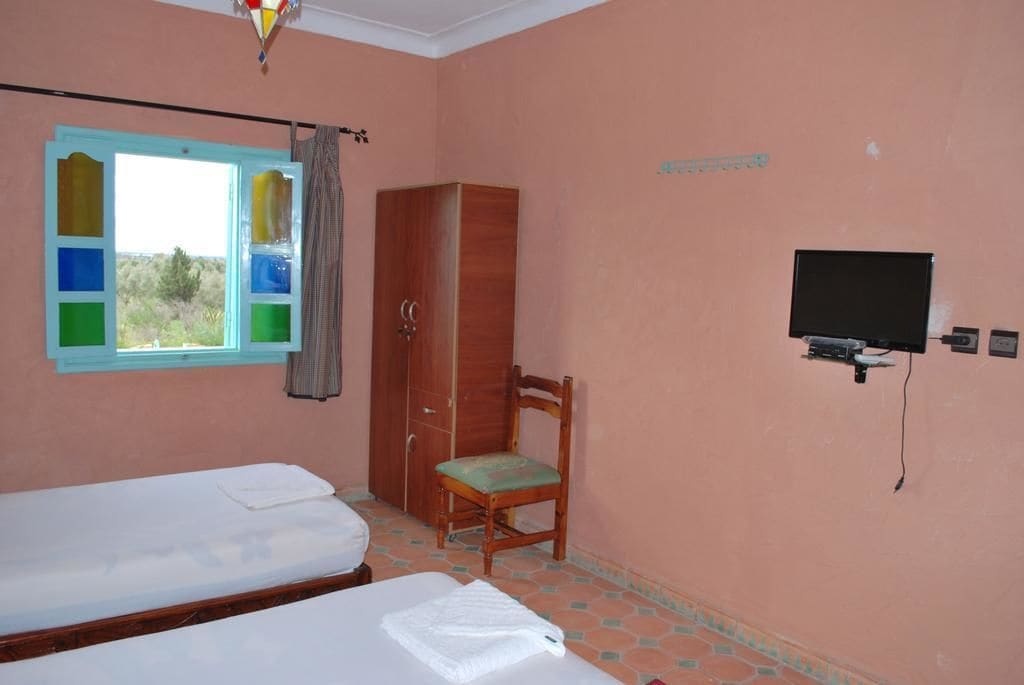 A room with two beds, a wardrobe, a chair, a television, and a window with a view of greenery.