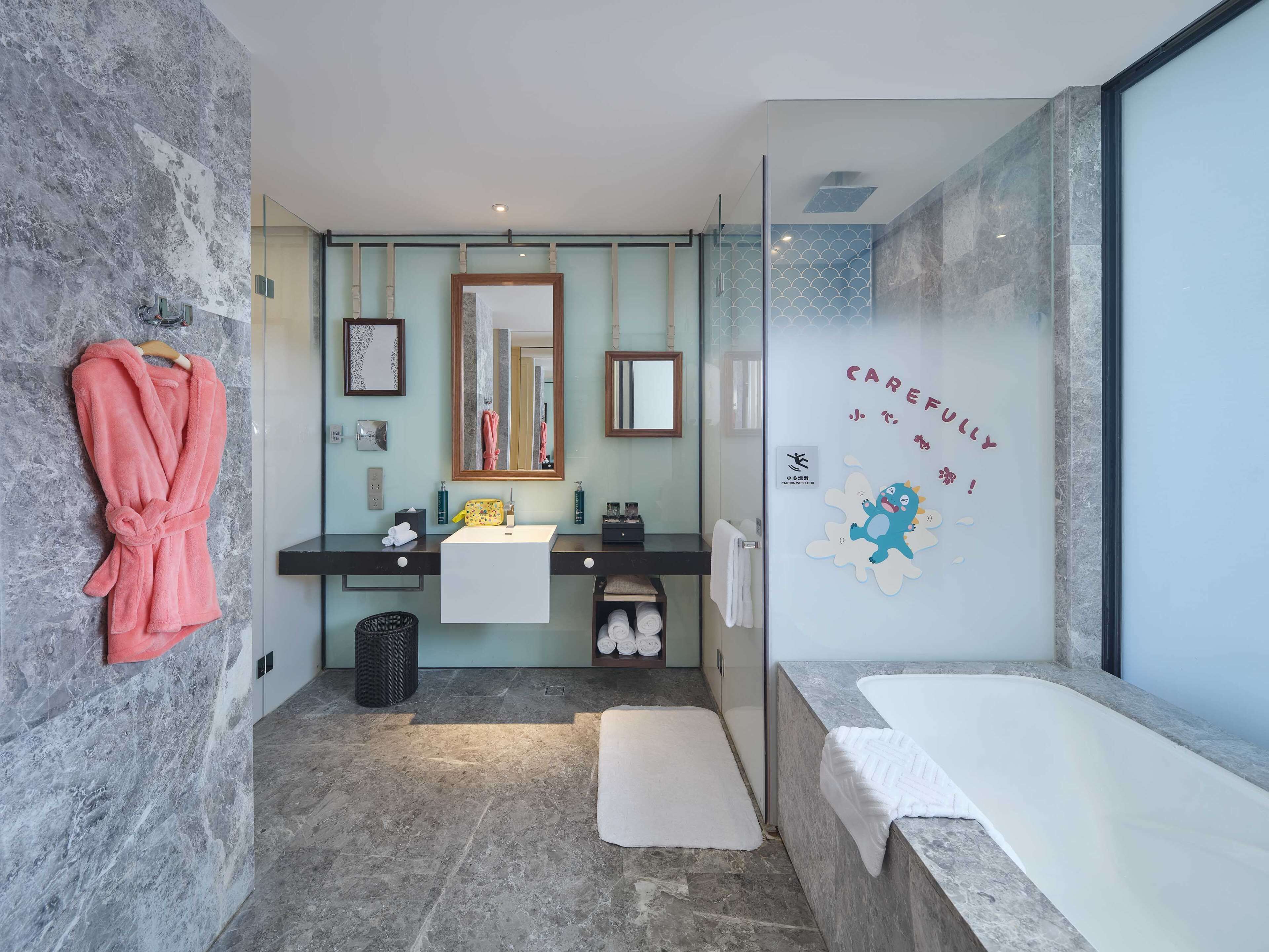 A modern bathroom with a stone wall, a large mirror, a sink, a bathtub, and a towel rack with a pink robe.