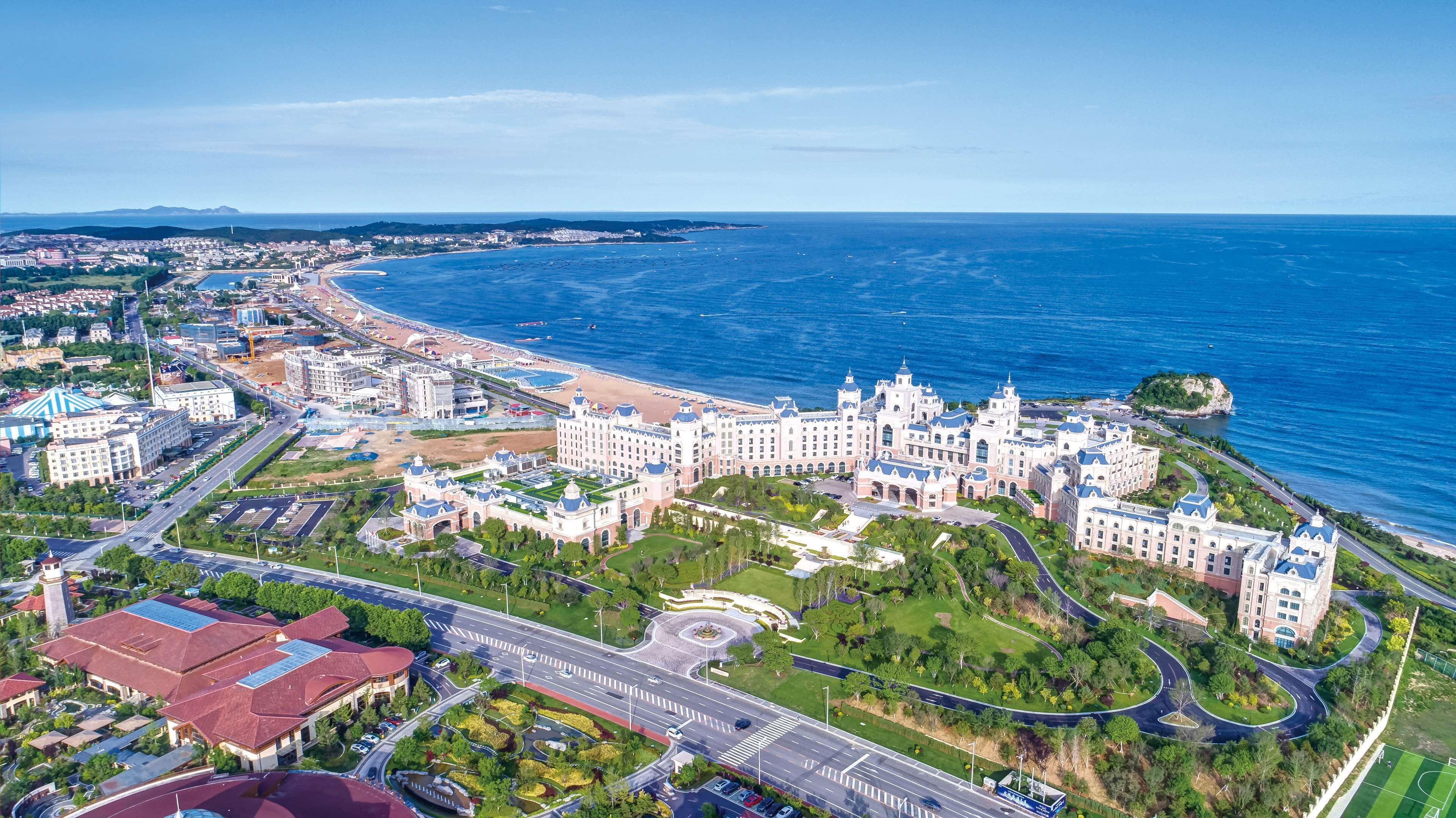 An aerial view of a coastal hotel with multiple buildings, a beachfront, and a well-maintained road network.