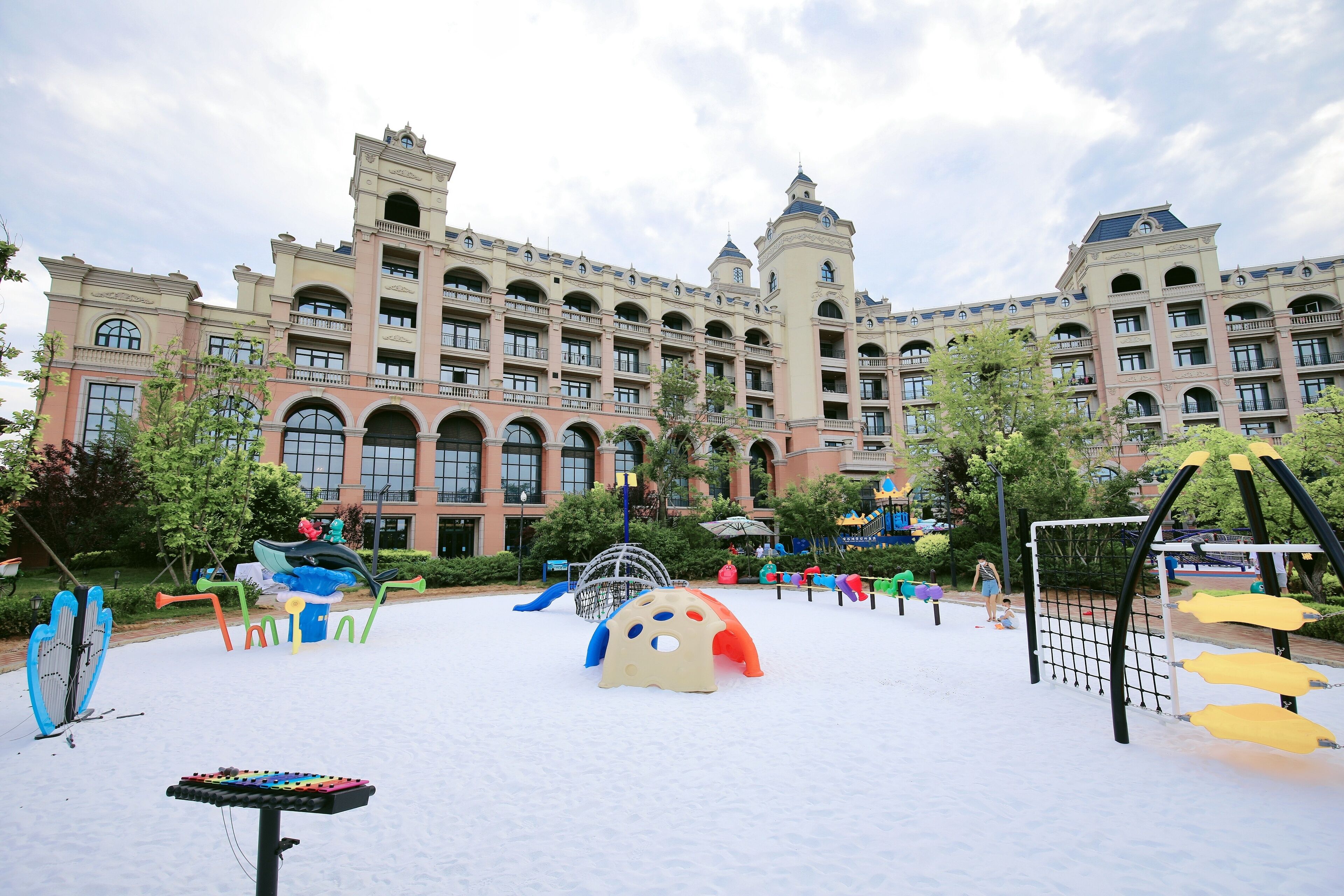 A playground area with various play structures, surrounded by a large building with multiple balconies and arched windows.