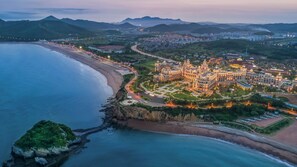 An aerial view of a coastal area with a large, illuminated building complex, a beach, and surrounding hills. - Hilton Dalian Golden Pebble Beach Resort (Dalian)