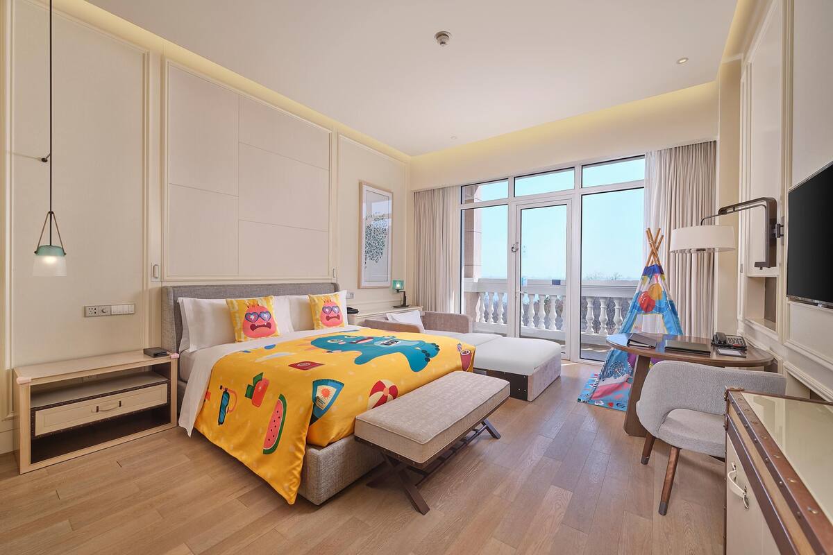 A hotel room with a bed, a desk, a chair, a balcony with a view, and a TV.