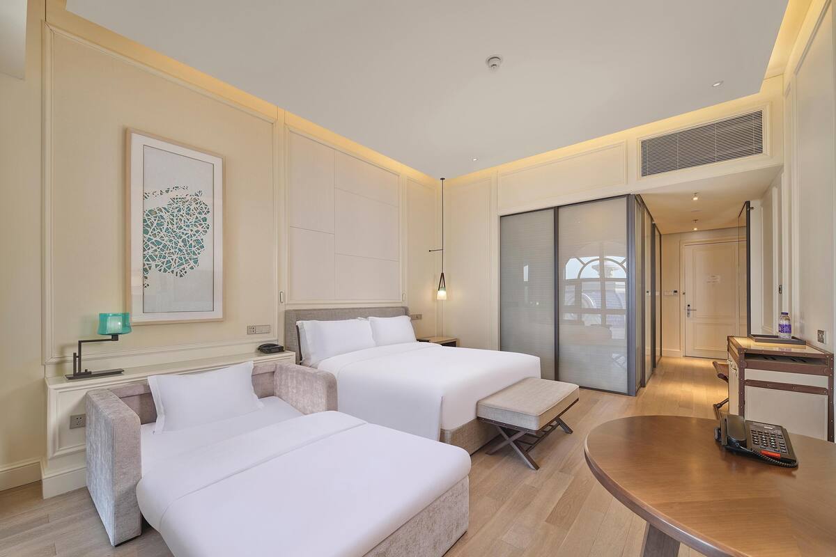 A hotel room with two beds, a desk, a chair, a table, and a large window.