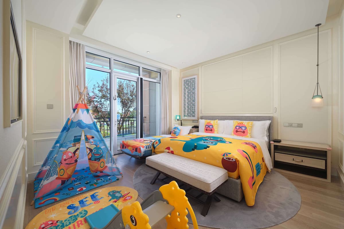 Een kindvriendelijke kamer met een bed, een bankje, een speelhoek met een tent en speelgoed, en een balkon met uitzicht.