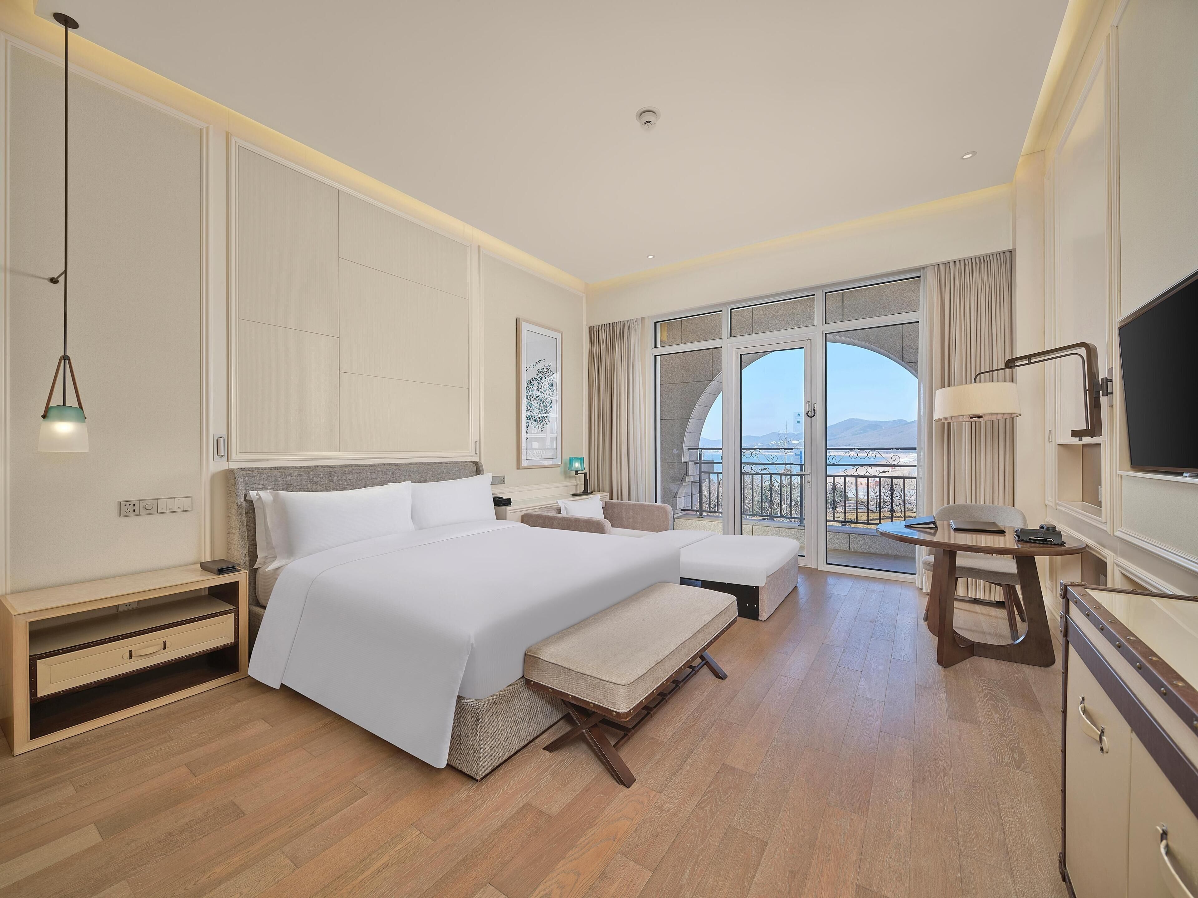 A hotel room with a large bed, a desk, a chair, a TV, and a balcony with a view.