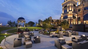 Un espace extérieur aménagé avec des meubles en osier et un kiosque, entouré d’une pelouse bien entretenue et éclairée, ainsi que d’un grand bâtiment à l’architecture classique.