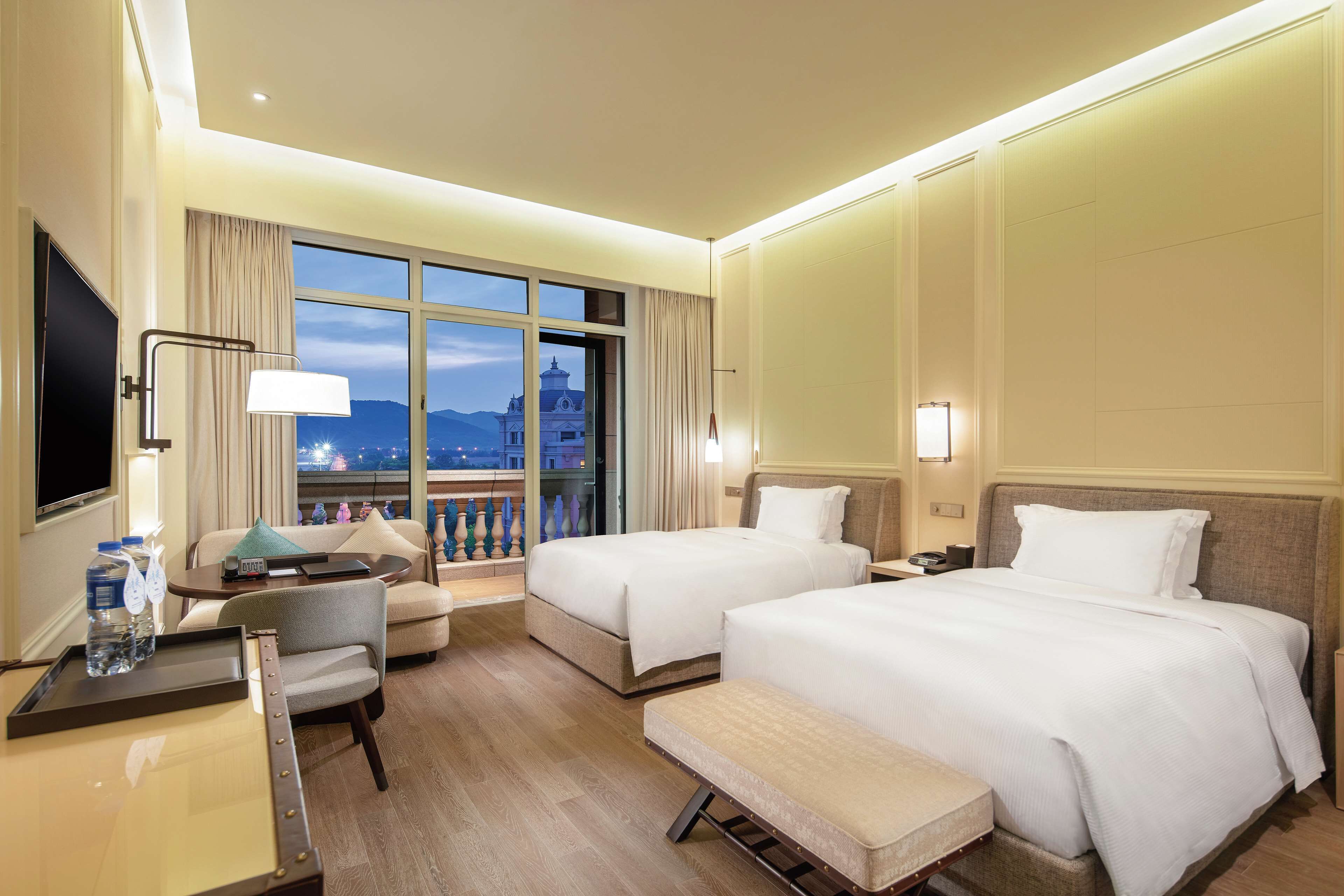 A hotel room with two beds, a desk with a chair, a TV, and a large window with a view of a building and mountains.