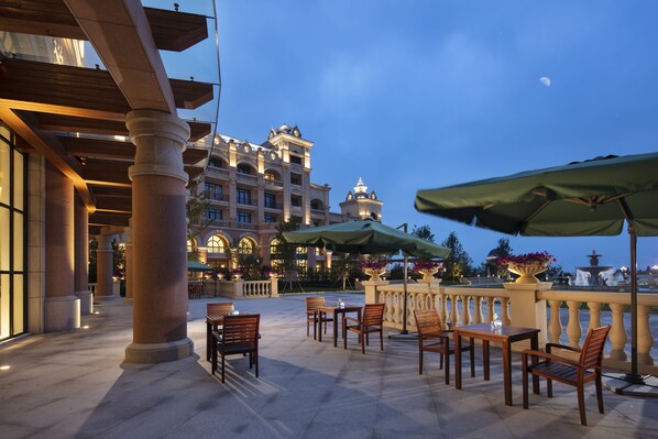 An outdoor patio with wooden tables and chairs, a fountain, and a large building in the background. - Hilton Dalian Golden Pebble Beach Resort (Dalian)