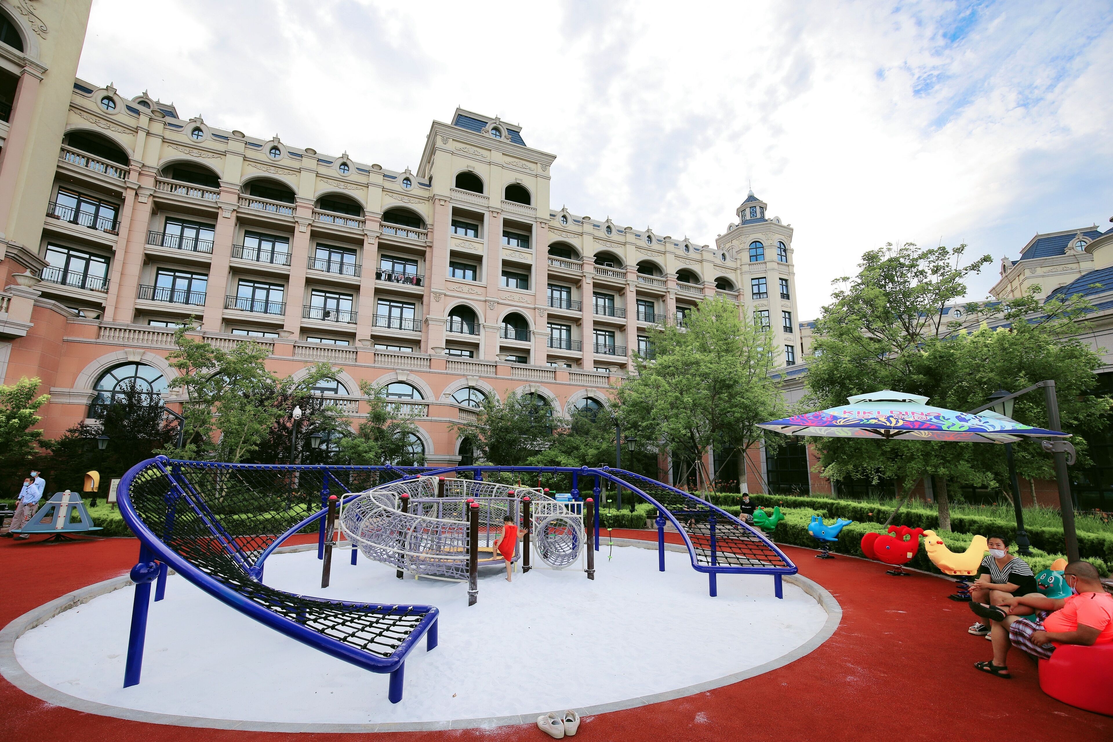 A playground with swings and climbing structures, surrounded by a red carpet area and trees.