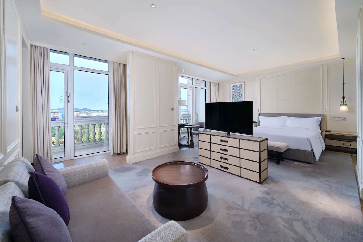 Een hotelkamer met een groot bed, een televisie, een bank, een salontafel en een balkon met gordijnen.