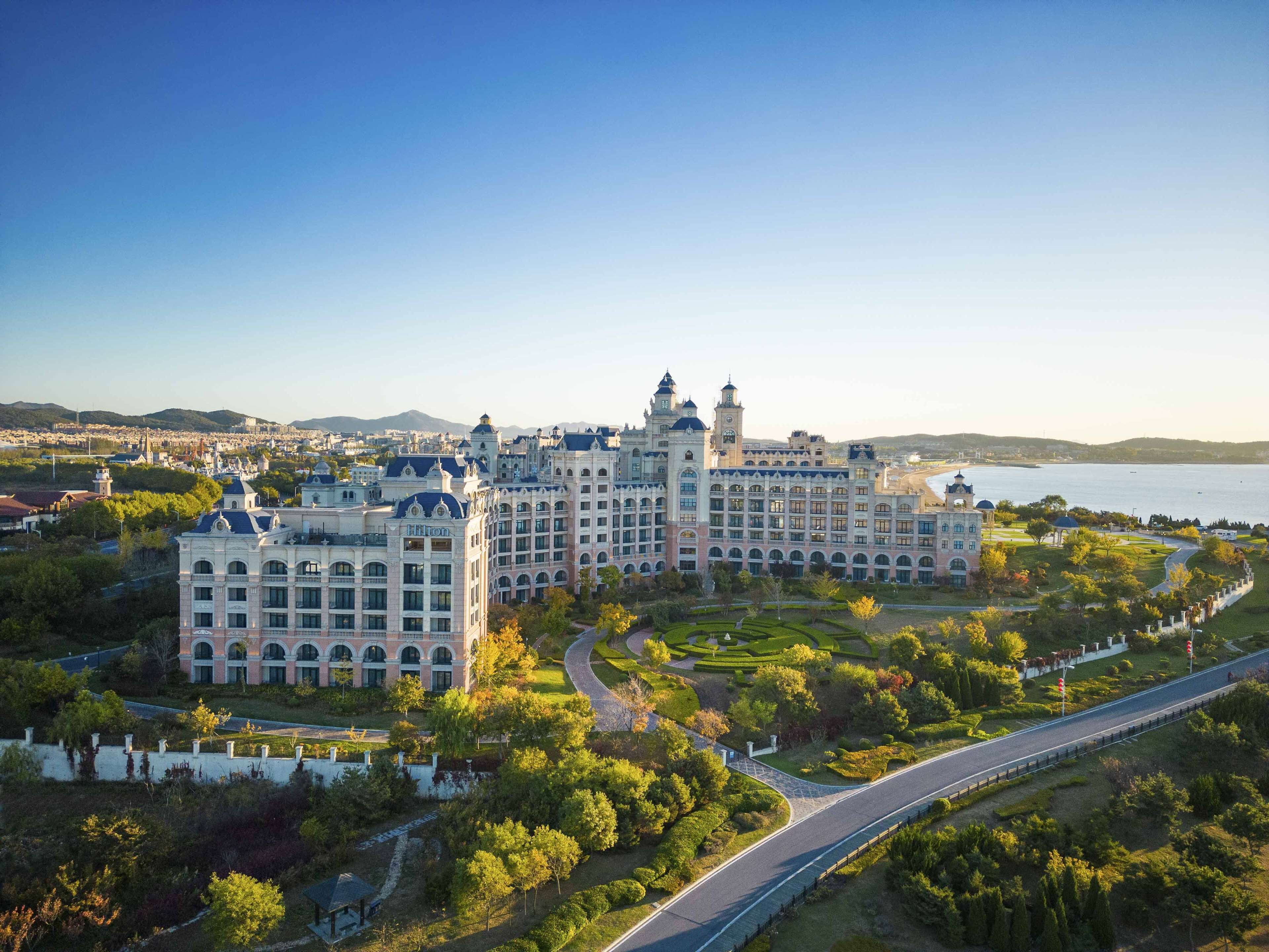 Un gran hotel histórico con múltiples torres y un patio central, rodeado de abundante vegetación y vistas al mar.