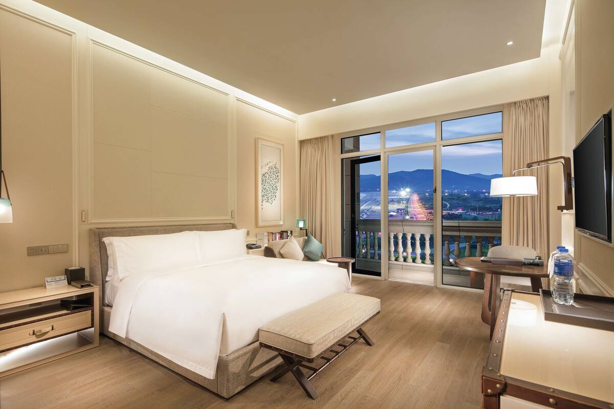 Hotelkamer met een groot bed, nachtkastjes, een bureau, een stoel, een televisie en een balkon met uitzicht op de stad.