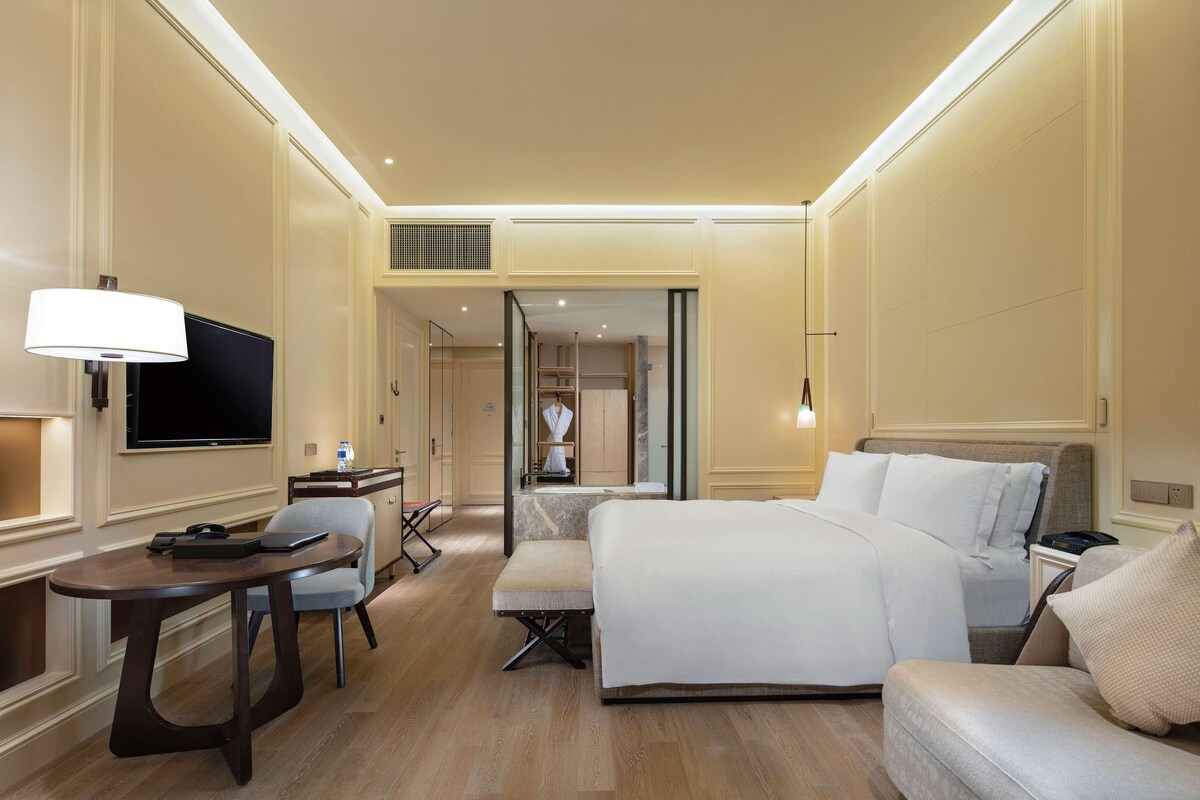 Habitación de hotel con cama, escritorio, silla, televisor y baño visible a través de una puerta abierta.