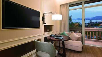 Una habitación de hotel con una gran televisión de pantalla plana, un escritorio con lámpara, un sofá y un balcón con vistas al mar y a las montañas.