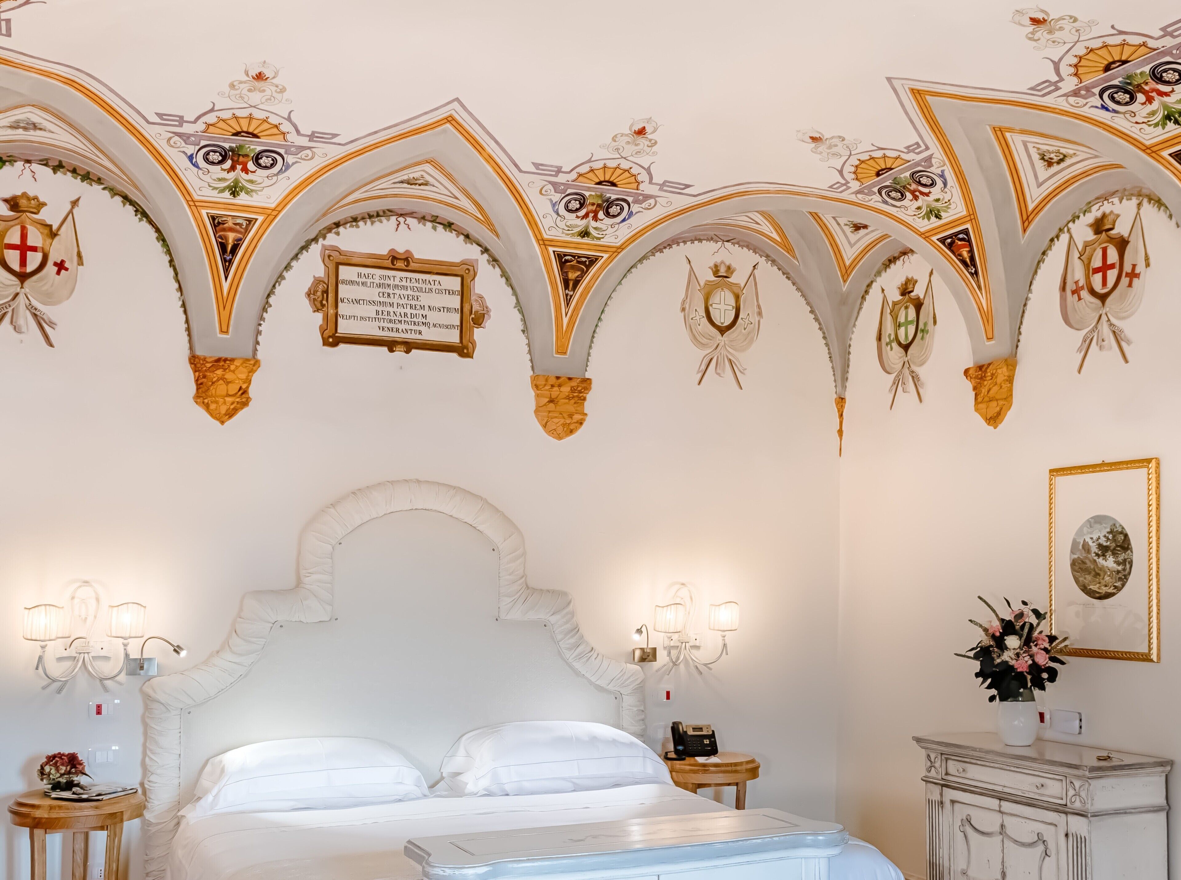 Habitación de hotel con una cama blanca, una araña y decoraciones ornamentales en el techo.
