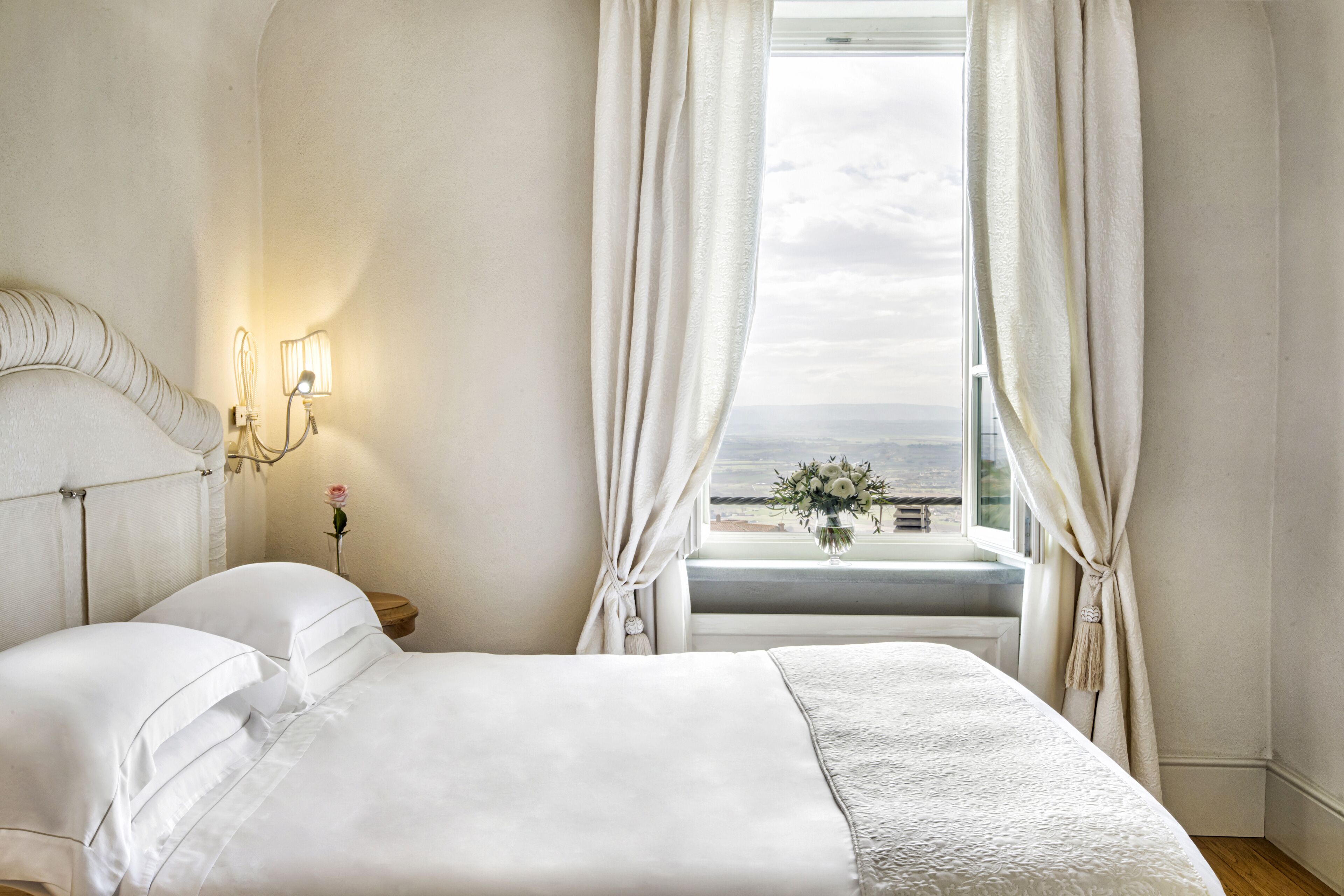 A bedroom with a bed, a nightstand with a lamp, a window with curtains, and a view of the landscape.