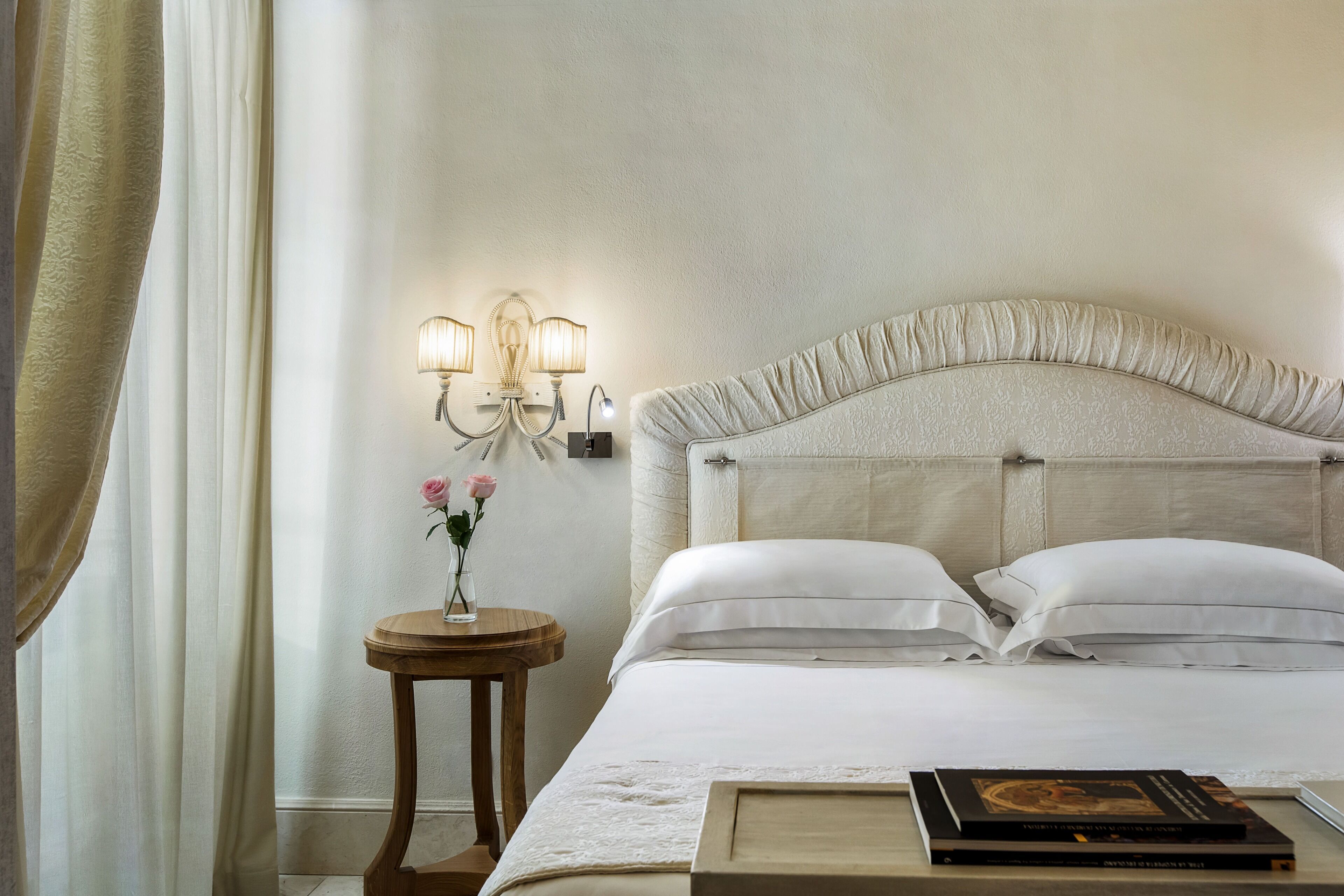 A neatly made bed with white linens, a bedside table with a vase of flowers, and a wall-mounted light fixture.