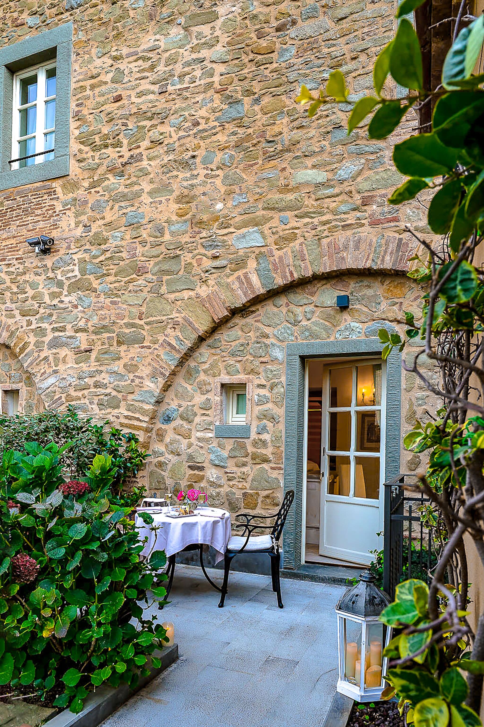 A stone building with a small patio area featuring a table and chairs, surrounded by greenery.