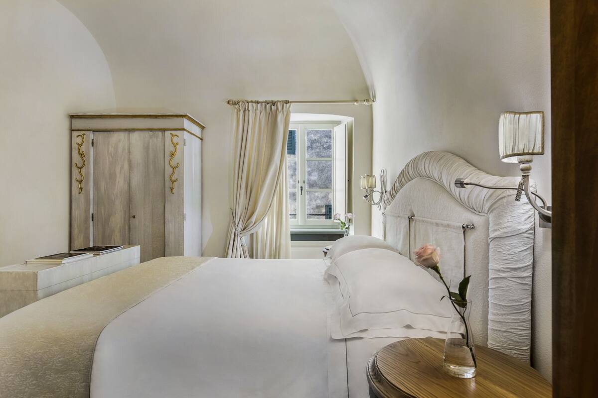 Een slaapkamer met een bed, nachtkastje, lamp, raam met gordijnen en uitzicht.