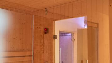 Ein Saunaraum mit Holzwänden und -bänken, einer Glastür zu einem Duschbereich und einem sichtbaren Bedienfeld für die Feuerung.