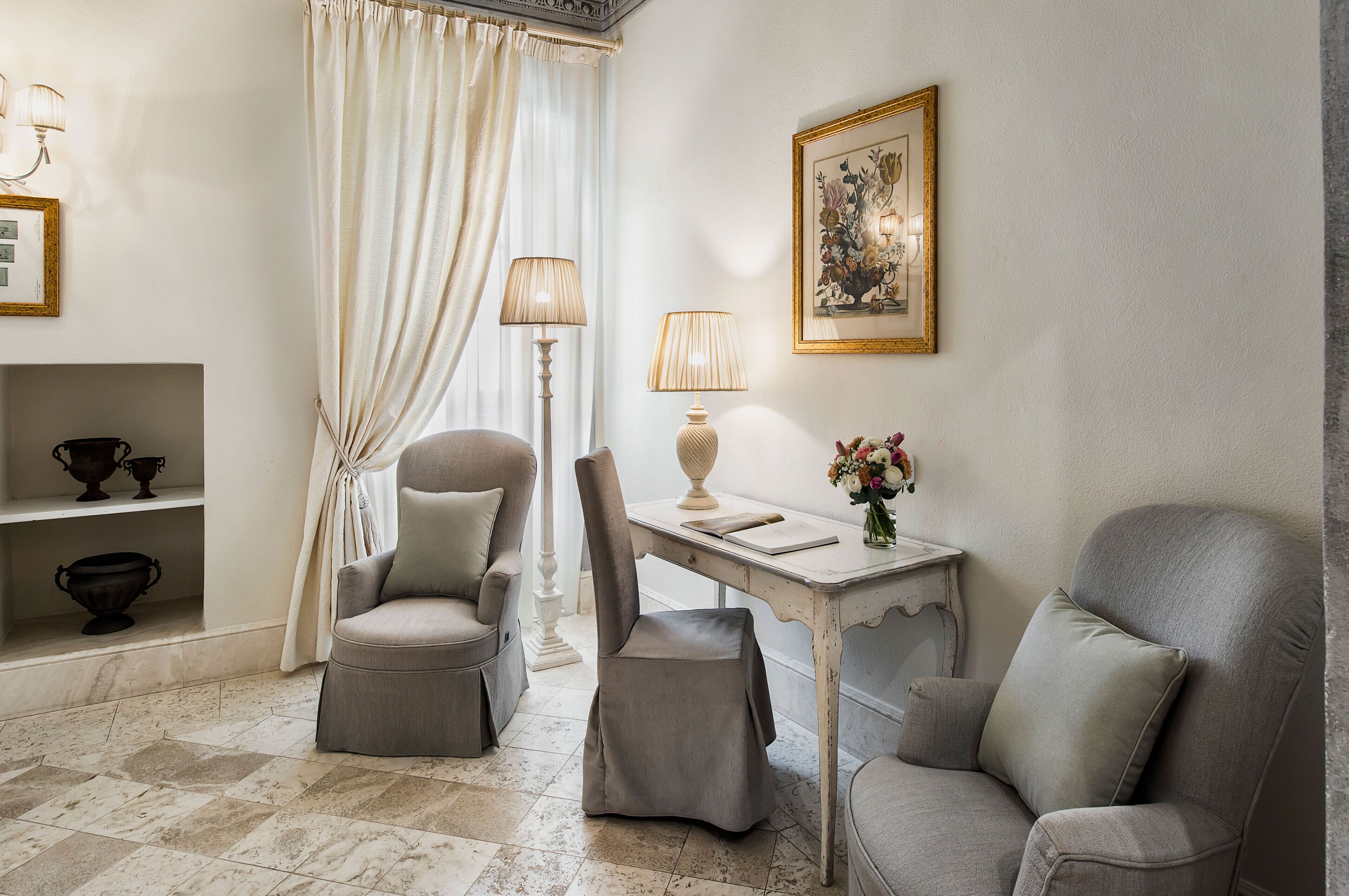 A room with a white table, two grey armchairs, a white floor lamp, and a framed floral painting on the wall.