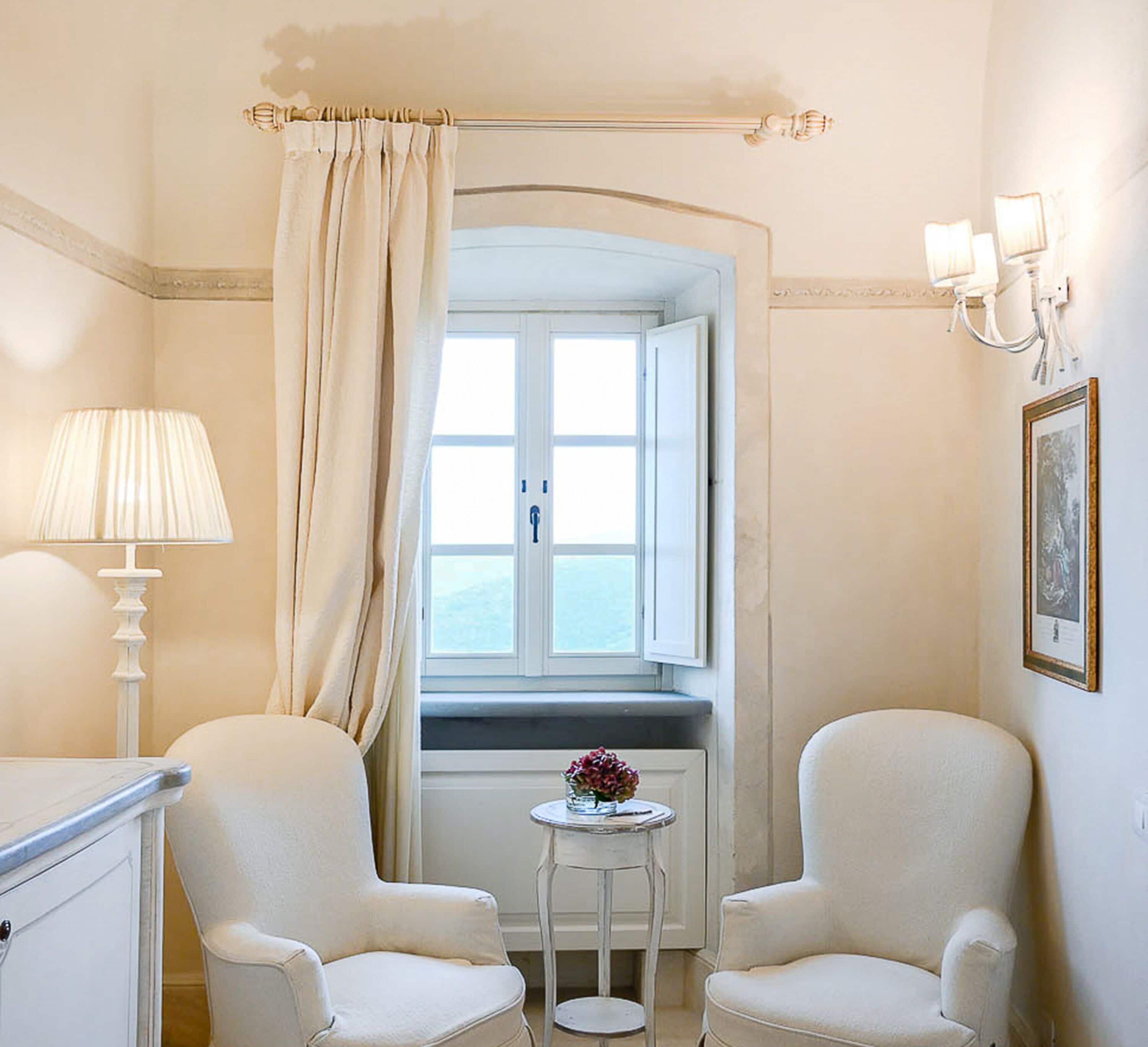 Une pièce comprenant deux fauteuils, une petite table avec un vase, une fenêtre donnant sur un beau paysage et une lampe.