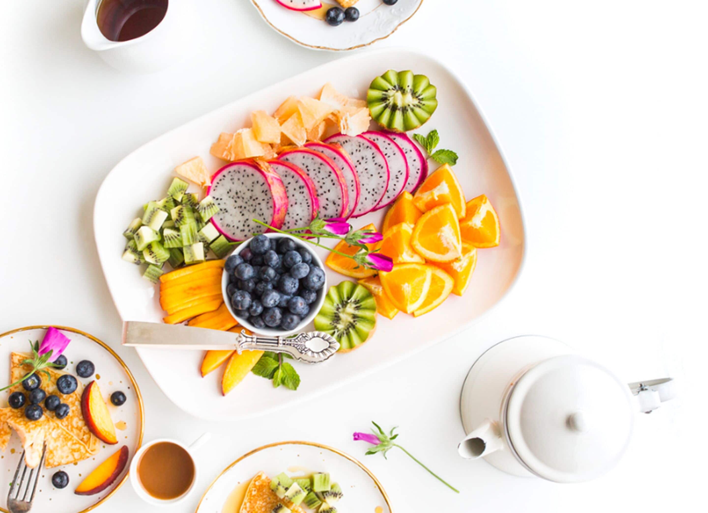 A white plate with sliced fruits and a bowl of blueberries, accompanied by a teapot and cups.