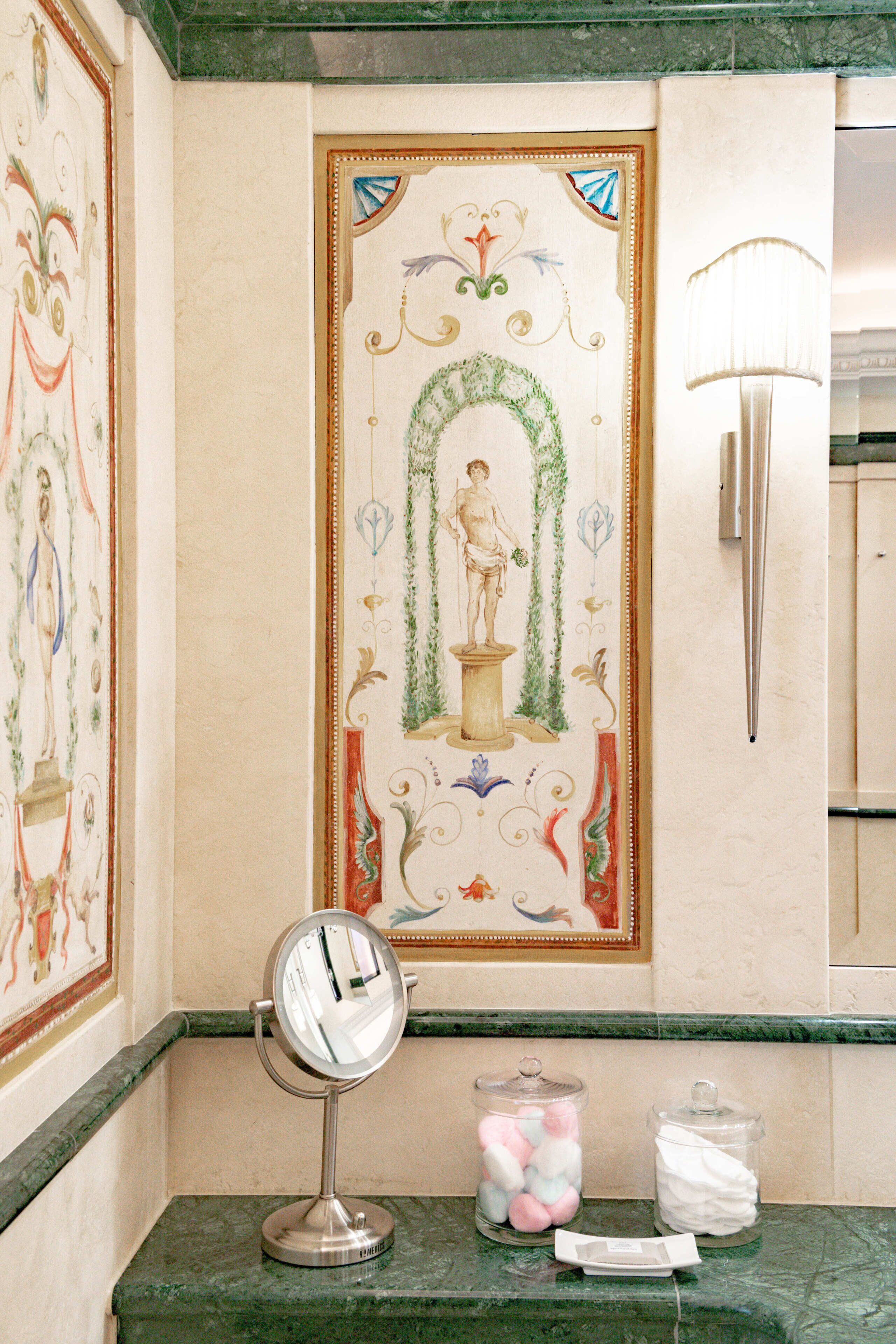 Een badkamer met een decoratief muurschildering, een make-up spiegel en een zeepdispenser.