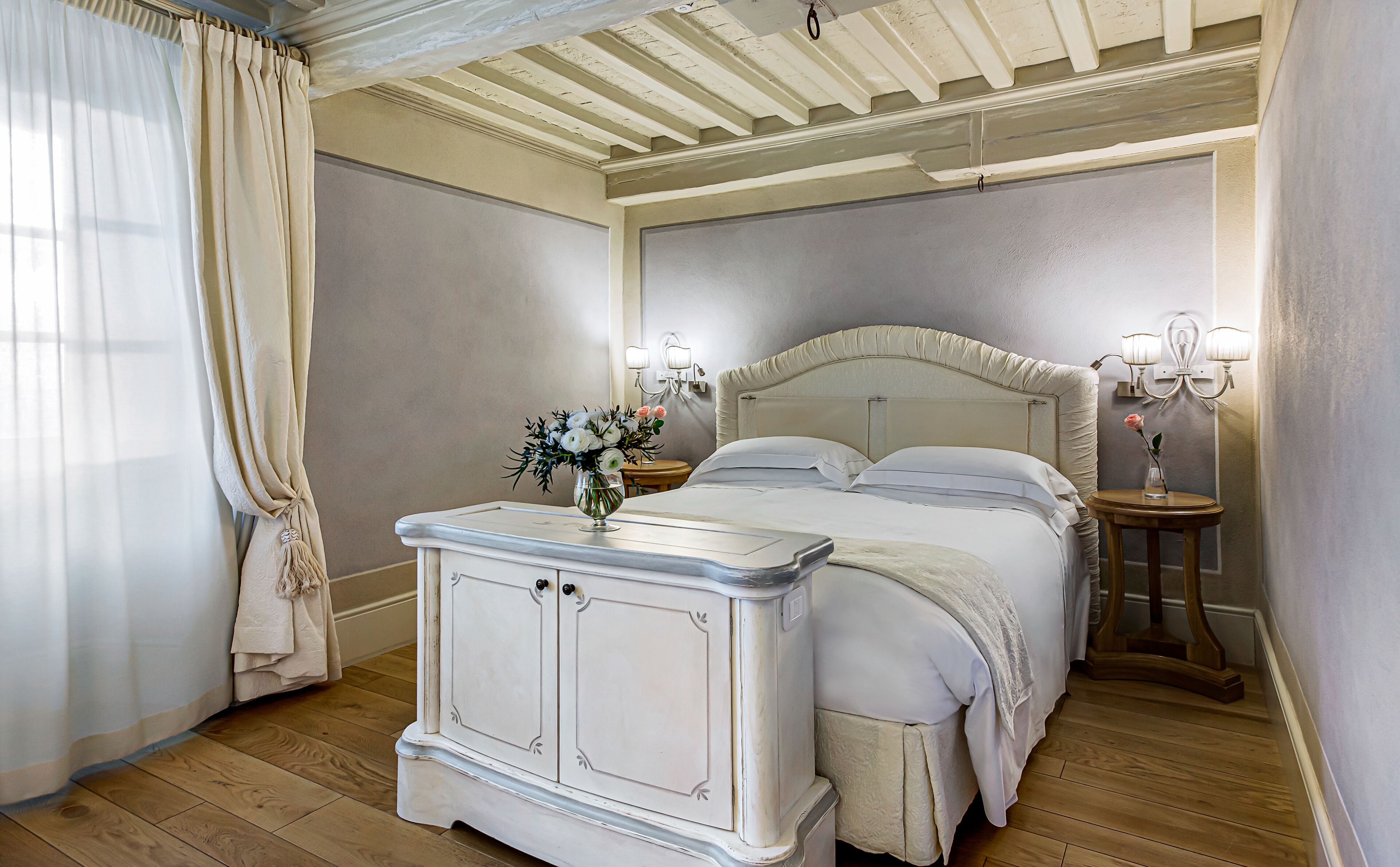 A bedroom with a large bed, a nightstand with a vase of flowers, and a wall-mounted lamp.
