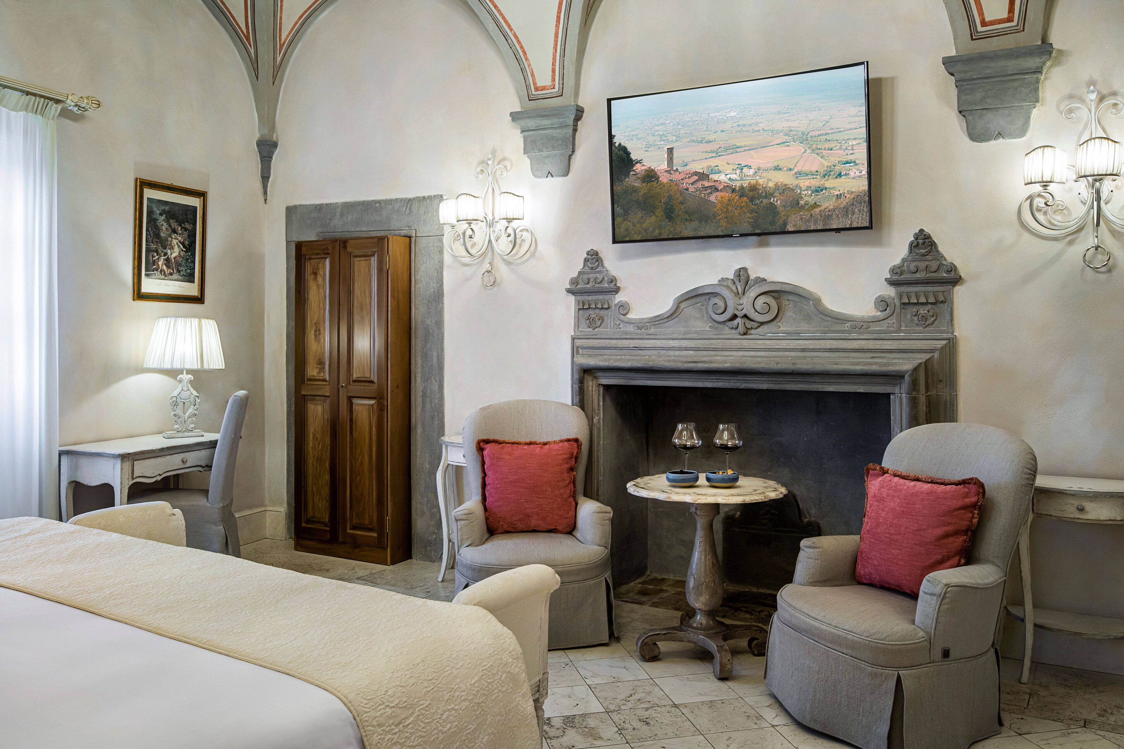 A hotel room with a bed, two armchairs, a small table with wine glasses, a fireplace, and a painting of a landscape.