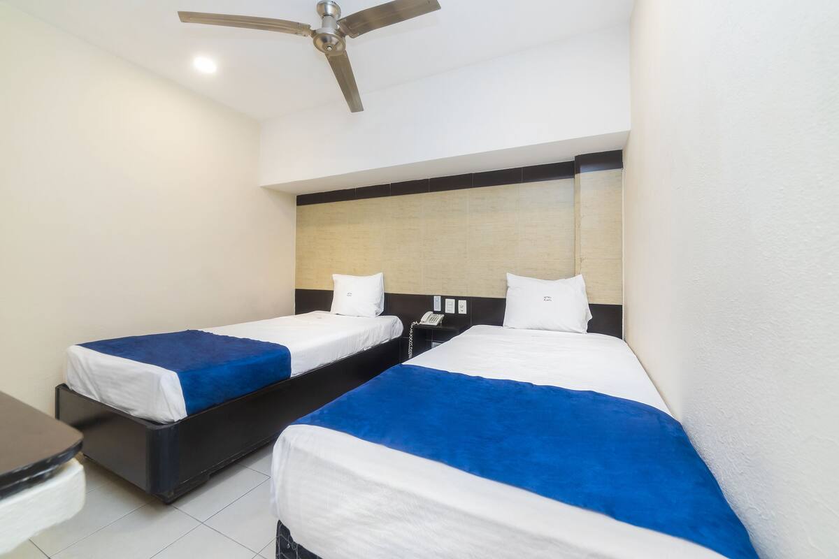A hotel room with two single beds, a desk, a chair, and a ceiling fan.